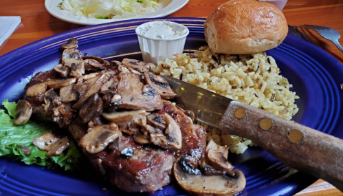 A plate featuring a steak topped with mushrooms, rice, a roll, and a small cup of sauce on a purple dish.