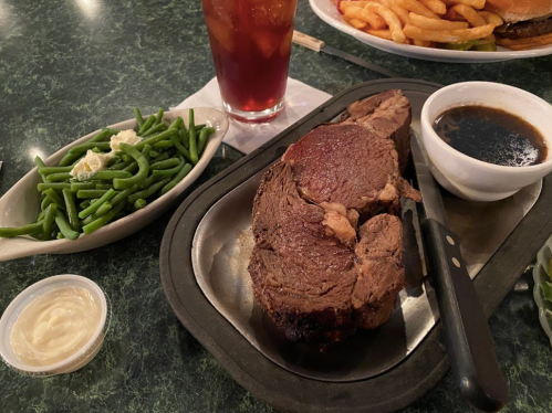 A plate with a large cut of beef, green beans, a small bowl of sauce, and a side of mayonnaise, with a drink in the background.