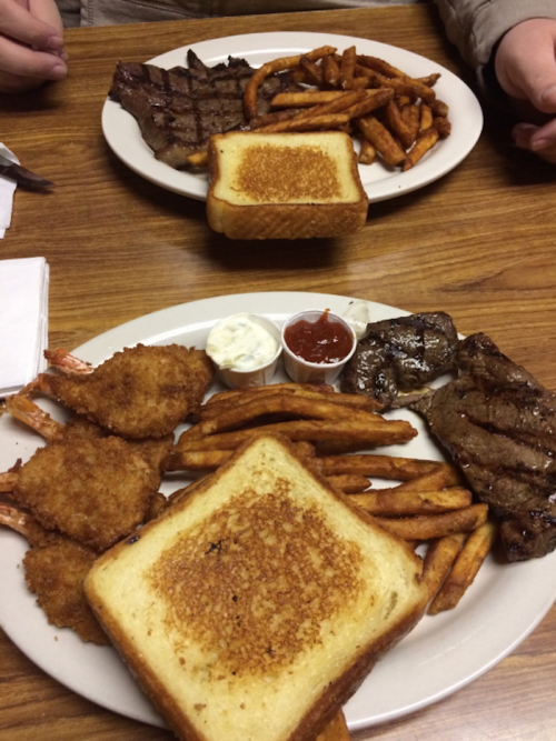 Two plates of food featuring steak, shrimp, fries, and toasted bread on a wooden table.