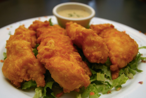 A plate of golden fried chicken tenders served on a bed of lettuce, accompanied by a small bowl of dipping sauce.