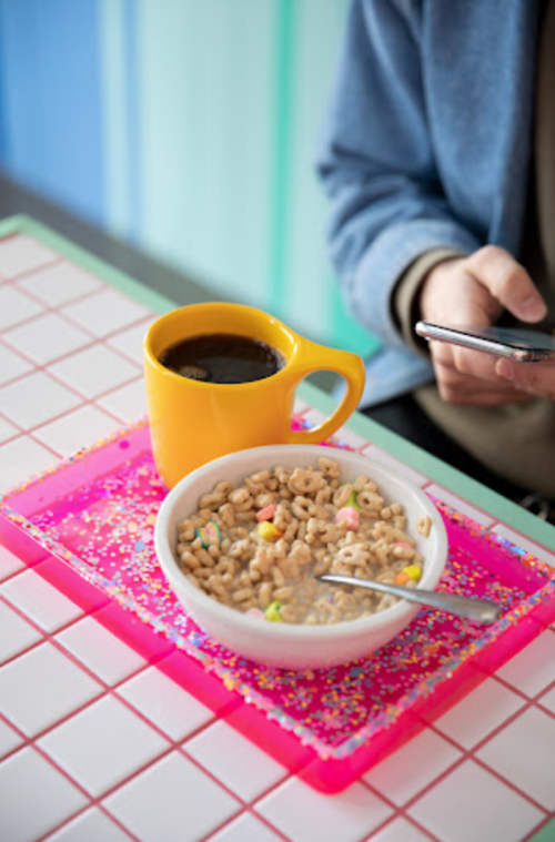 A bowl of cereal with colorful pieces next to a yellow mug of coffee on a bright, patterned tray. A hand holds a phone.