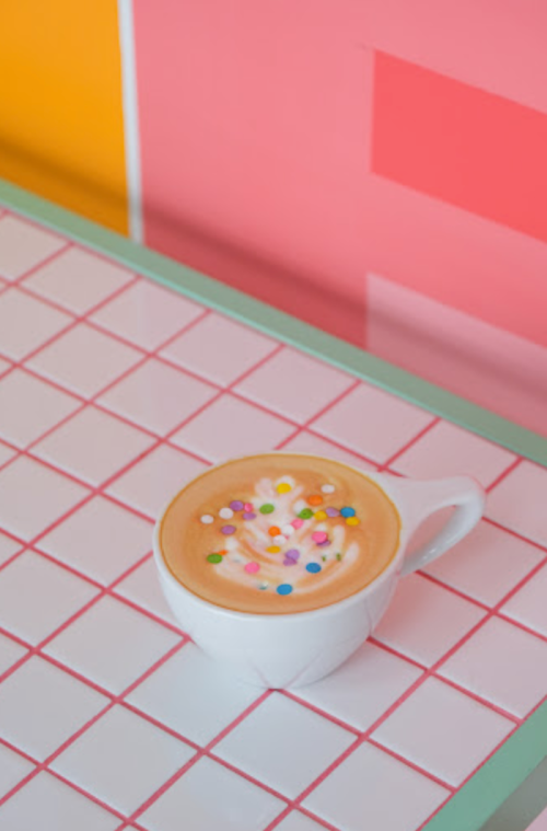A cup of coffee topped with colorful sprinkles on a tiled table with a vibrant background.