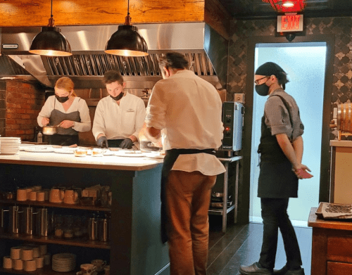 Four chefs in a restaurant kitchen prepare dishes, wearing masks and working at a counter with various kitchen tools.
