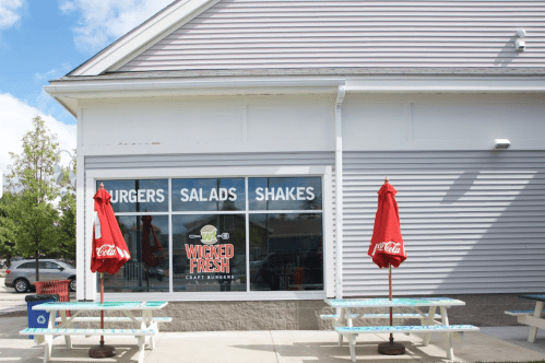 Exterior of Wicked Fresh restaurant featuring a sign for burgers, salads, and shakes, with red umbrellas over picnic tables.