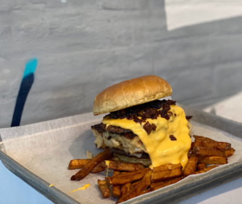 A loaded burger with melted cheese and crispy toppings, served with a side of golden fries on a metal tray.