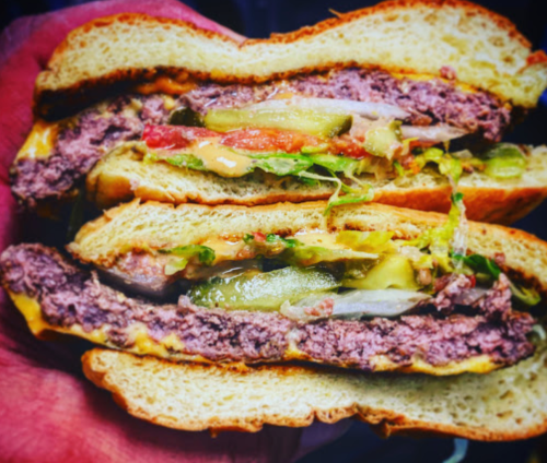 A close-up of a juicy burger with lettuce, tomato, pickles, and cheese, held between two slices of bread.