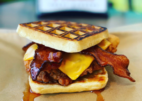 A crispy chicken sandwich with bacon and cheese, served between two golden waffles drizzled with syrup.