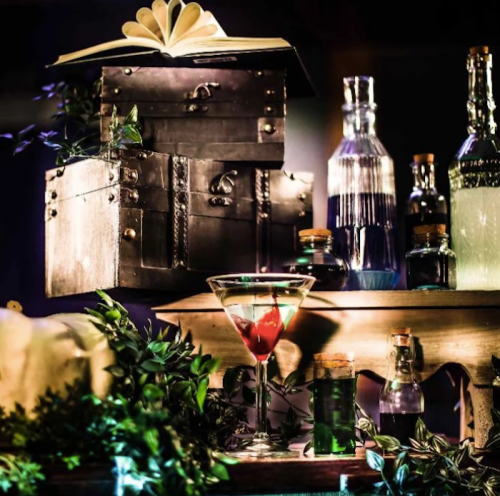 A stylish display of vintage bottles, a cocktail with a cherry, and an open book, surrounded by greenery and soft lighting.