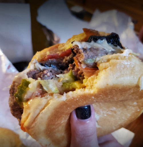 A hand holding a half-eaten burger with layers of meat, bacon, lettuce, and toppings on a soft bun.