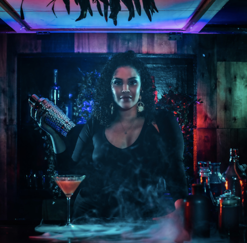 A bartender with curly hair stands behind a bar, holding a shaker, surrounded by colorful lights and smoke.