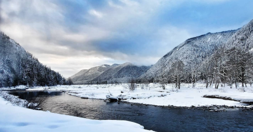 A serene winter landscape featuring a river surrounded by snow-covered mountains and trees under a cloudy sky.