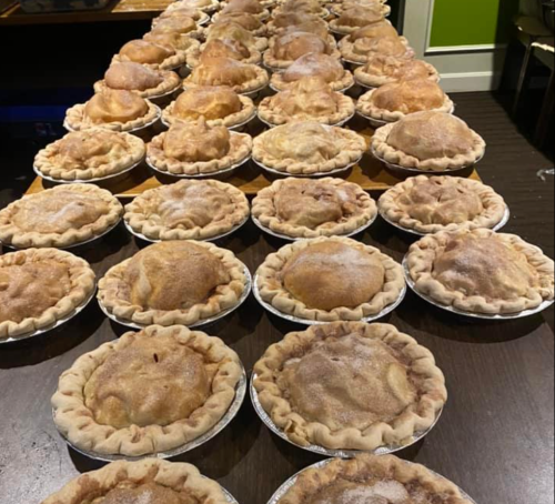 A long table lined with freshly baked apple pies in silver foil pans, each topped with a sprinkle of sugar.