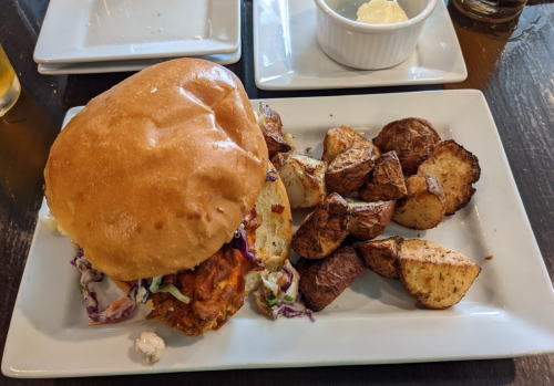 A plate with a sandwich filled with pulled meat and coleslaw, accompanied by seasoned potato wedges and a small bowl of sauce.