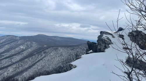 A snowy mountain landscape with rocky outcrops and distant hills under a cloudy sky.