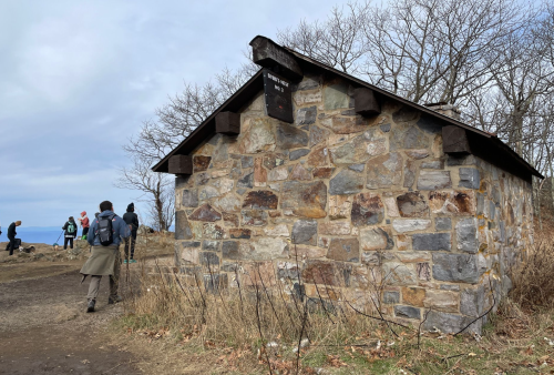A stone building with a sloped roof, surrounded by bare trees and people walking nearby on a cloudy day.