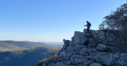 A hiker stands on a rocky outcrop, overlooking a vast mountain landscape under a clear blue sky.