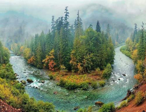 A serene river winding through lush green and autumn-colored trees, surrounded by misty mountains.