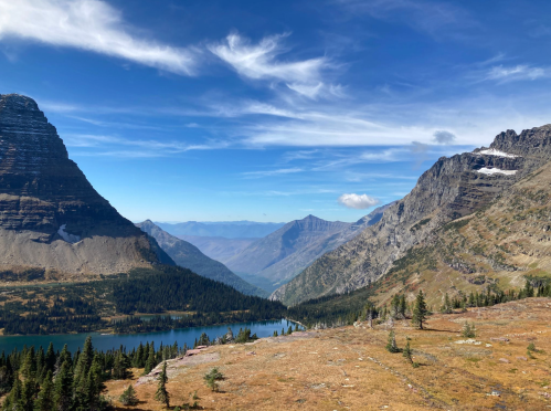 A scenic view of mountains and a lake under a blue sky with wispy clouds, surrounded by trees and rocky terrain.