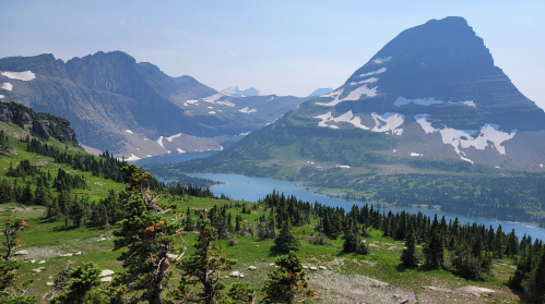 A scenic view of mountains and a lake, surrounded by lush greenery and patches of snow under a clear blue sky.