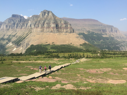 A wooden path leads through a green landscape with mountains in the background and three hikers walking ahead.