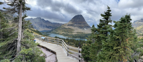 A scenic view of a mountain and lake, framed by trees and a wooden walkway under a cloudy sky.