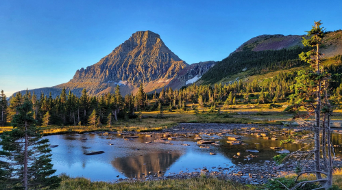 A serene mountain landscape with a peak reflecting in a calm pond, surrounded by trees and clear blue skies.