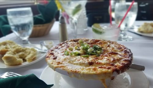A bowl of cheesy baked dish topped with green onions, surrounded by drinks and bread rolls on a dining table.