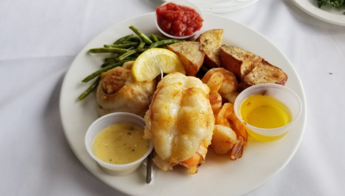 A plate of seafood featuring shrimp, lobster, green beans, seasoned potatoes, and dipping sauces.