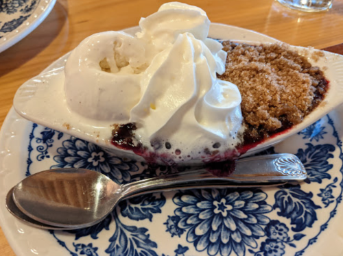 A dessert dish with ice cream, whipped cream, and a crumbly topping on a floral plate, accompanied by a spoon.