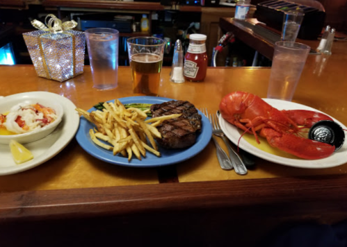 A table setting with a lobster, steak with fries, and a side dish, accompanied by drinks and condiments.