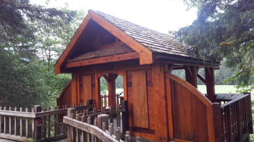 A wooden structure with a sloped roof, surrounded by trees and a wooden fence, set in a natural landscape.