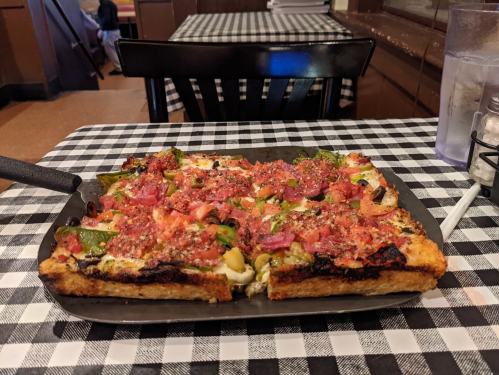 A rectangular pizza topped with vegetables and herbs on a black plate, set on a checkered tablecloth.