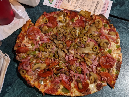 A large pizza topped with pepperoni, ham, green peppers, olives, and mushrooms on a dark table.