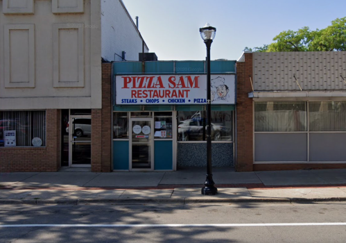 Front view of Pizza Sam restaurant, featuring a sign with menu items and a street lamp nearby.