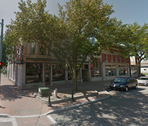 A street view of a corner building with a tree, featuring a restaurant sign and parked cars on a sunny day.