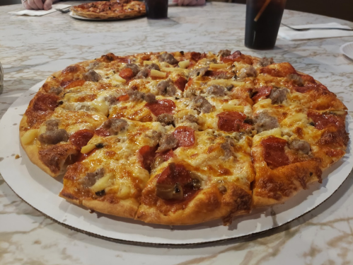 A close-up of a pizza topped with pepperoni, sausage, and cheese on a marble table, with drinks in the background.