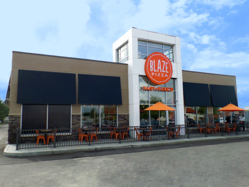 Exterior of Blaze Pizza restaurant featuring large windows, orange umbrellas, and a modern design.