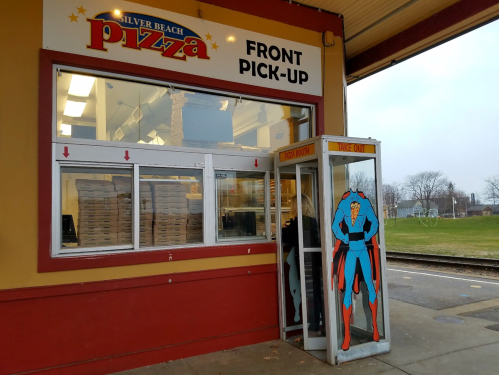 A pizza pickup window with a superhero-themed phone booth beside it at Silver Beach Pizza.