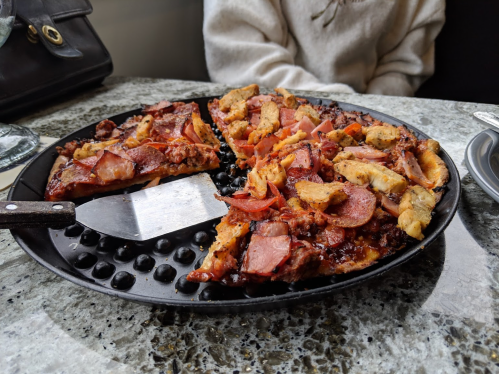 A close-up of a pizza on a round pan, topped with meats and vegetables, with a pizza cutter beside it.