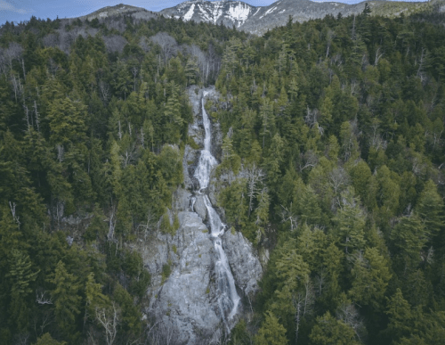 Aerial view of a cascading waterfall surrounded by lush green trees and distant snow-capped mountains.