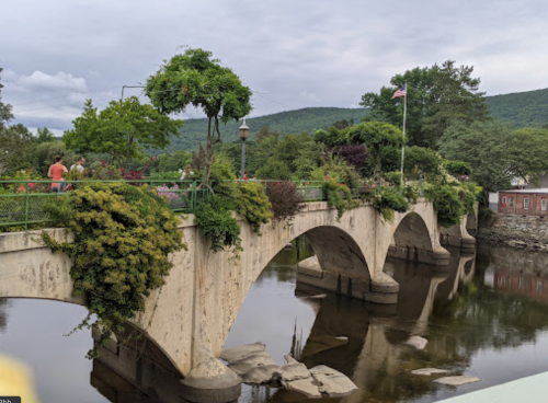 A stone bridge adorned with greenery spans a calm river, surrounded by trees and hills under a cloudy sky.