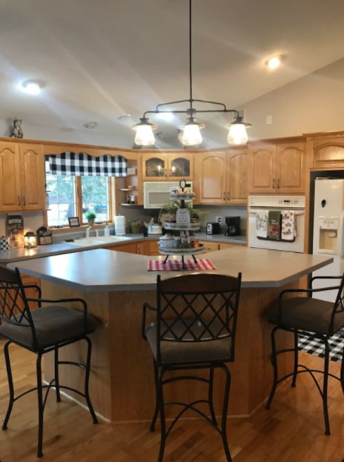 A cozy kitchen with wooden cabinets, a central island, bar stools, and warm lighting. Decor includes checkered patterns.