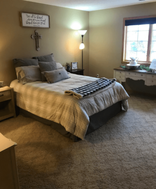 Cozy bedroom with a bed, decorative pillows, a lamp, and a desk by a window, featuring warm, neutral tones.