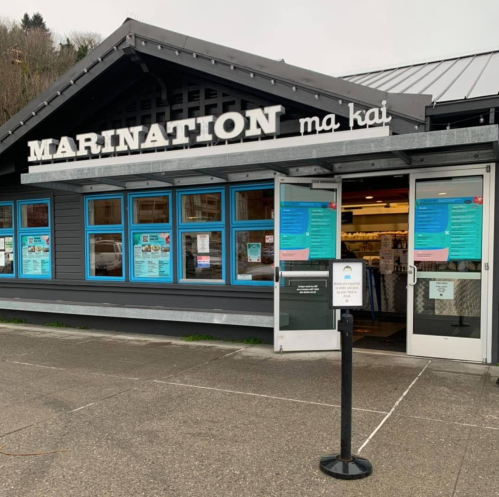 Exterior of Marination Ma Kai restaurant, featuring a sign and large windows with blue accents.