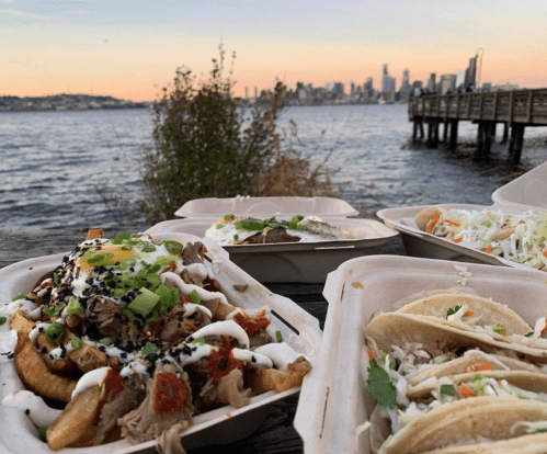 A scenic view of food containers with tacos and loaded fries, overlooking a waterfront and city skyline at sunset.