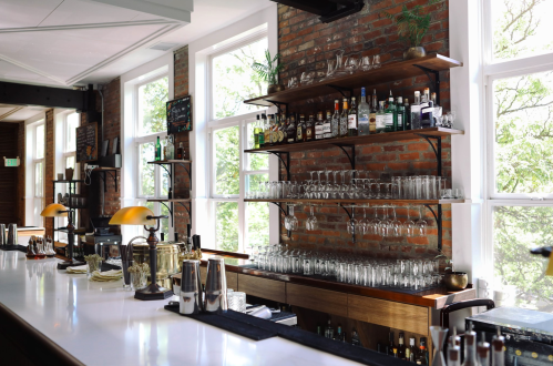 A stylish bar with a wooden counter, shelves of liquor, and large windows letting in natural light.