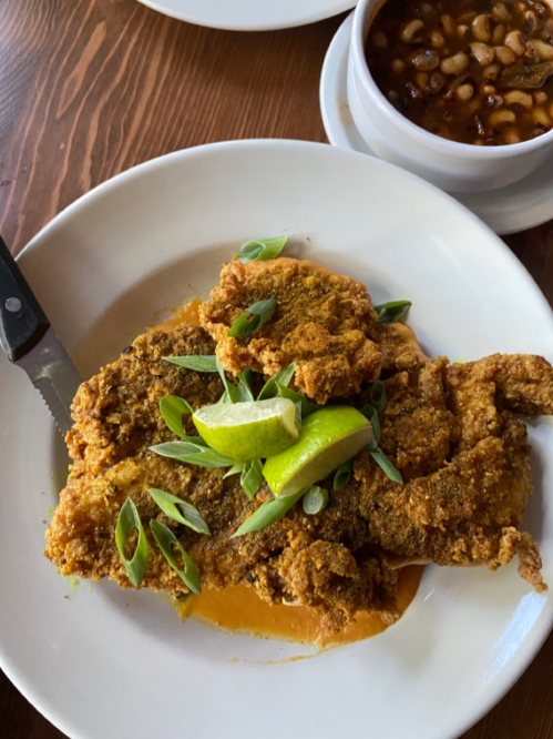 A plate of fried fish topped with green onions and lime, served with a bowl of black-eyed peas in the background.