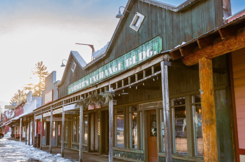 Historic wooden buildings line a snowy street, featuring a sign for the "Farmer's Exchange Bldg." in soft sunlight.