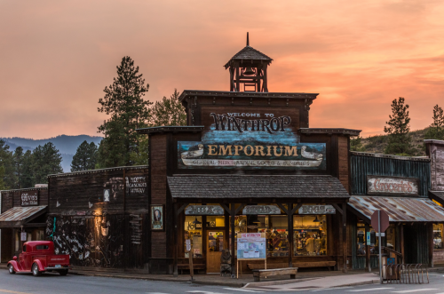 A rustic wooden building with a sign reading "Welcome to Winthrop Emporium" at sunset, surrounded by trees and mountains.