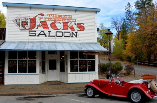 A vintage red car parked in front of "Three-Fingered Jack's Saloon," a rustic building with a colorful sign.